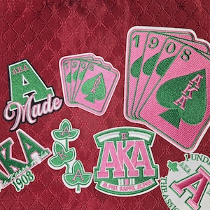 May include: A collection of embroidered patches featuring pink and green designs. The patches include text such as AKA, A Made, 1908, and Alpha Kappa Alpha, alongside imagery of playing cards with spade symbols and ivy leaves on a textured red background.