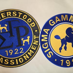 May include: Two circular patches with embroidered designs. One features the text "I UNDERSTOOD THE ASSIGNMENT" and "1922" with poodle silhouettes. The other says "SIGMA GAMMA RHO" and "1922" with a poodle silhouette. The patches have blue, yellow, and black colors.