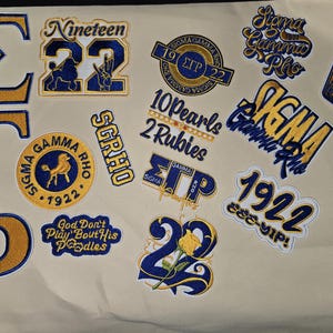 May include: Embroidered patches in gold and navy blue, featuring Greek letters, numbers, and phrases like "Sigma Gamma Rho" and "1922". The patches are on a cream-colored fabric, ideal for jackets or bags.
