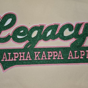 May include: Embroidered patch with the word "Legacy" in a cursive font above "Alpha Kappa Alpha". The text is green with a pink outline and white accents, set against a tan background.