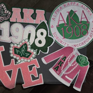 May include: A collection of pink, green, and white embroidered patches and appliques. Includes the letters "AKA", the number "1908", a jacket, and a heart-shaped design. The patches are for Alpha Kappa Alpha Sorority.