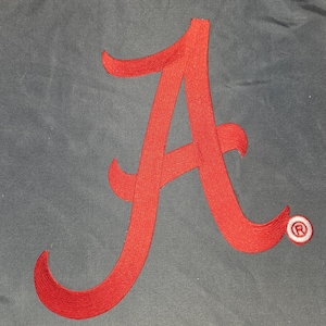 May include: A large, embroidered red letter "A" on a gray background. The letter has a stylized, cursive design. A small, circular emblem with a white "R" inside a red circle is in the lower right corner. The design is likely for apparel or accessories.