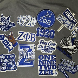 May include: A collection of embroidered patches in blue and white, featuring various designs including text, a dove, and a hand gesture. The patches display phrases like "Zeta 20" and "Finer Way".