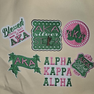 May include: A collection of embroidered patches in shades of green, pink, and white. The patches feature the letters "AKA" and phrases like "Blessed Pretty Girl of AKA" and "Alpha Kappa Alpha".