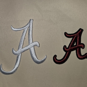 May include: Two embroidered patches featuring the letter A in a script font. One patch is white with a silver sheen, and the other patch is dark crimson red. Both patches have a raised, textured stitch pattern and are displayed on a tan fabric.
