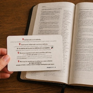 Puede incluir: Un libro abierto con texto, junto a una tarjeta pequeña con texto en español. Una taza de café está sobre un posavasos en el fondo. El texto de la tarjeta incluye las palabras "Esfuérzate y sé valiente."