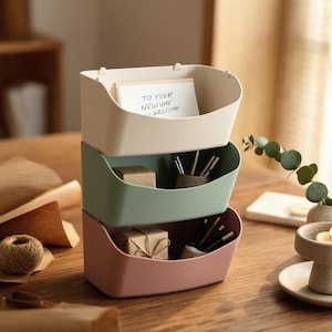 May include: A stack of three wall-mountable storage bins in cream, sage green, and dusty rose. The top bin holds a white card with the text "TO YOUR NEW HOME - WELCOME". The middle and bottom bins contain small wrapped packages and pens.