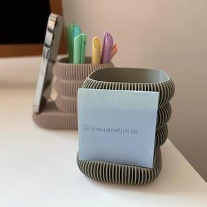 Op de afbeelding: Twee geribbelde, cilindervormige bureau-organizers in gedempt taupe en olijfgroen. De taupekleurige organizer bevat een smartphone en diverse pastelkleurige pennen. Een lichtblauw memoblaadje met de tekst @SHQNAVIIRLUR.SD zit op de olijfgroene organizer.