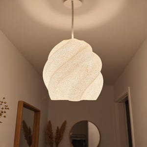 May include: A white pendant light fitting with a textured, spiralling, twisted shape. The lamp is suspended from the ceiling by a single cord and emits a soft, warm glow, illuminating the surrounding space with a diffused light pattern.