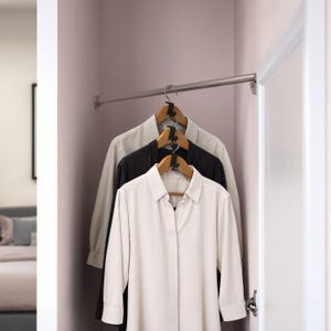 May include: Three button-down shirts hanging in a wardrobe. The shirts are light beige, black, and white. The shirts are on wooden hangers with black hooks, suspended from a silver rail. The wardrobe walls are a light purple colour.