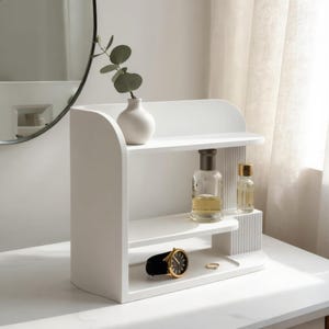 Pode incluir: Uma prateleira organizadora branca de vários níveis com laterais curvas e um painel canelado. A prateleira superior contém um pequeno vaso branco com uma planta verde, enquanto os níveis inferiores exibem um frasco de vidro e um relógio.