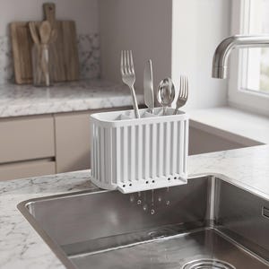 May include: A white rectangular cutlery drainer with vertical ribbed sides and multiple internal compartments. The drainer is positioned on the edge of a stainless steel sink, holding two silver forks, a knife, and a spoon, with water droplets.