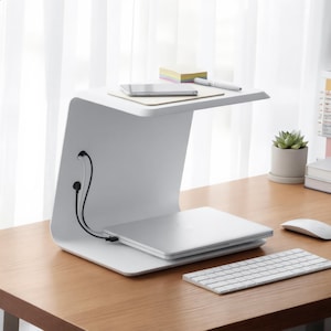 Könnte beinhalten: Ein weißer, C-förmiger Laptopständer mit zweistufigem Design. Die obere Ebene trägt ein Smartphone, einen Stift und einen Stapel Haftnotizen. Die untere Ebene stützt einen geschlossenen silbernen Laptop, der mit einem schwarzen Kabel verbunden ist.
