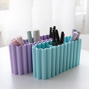 Op de afbeelding: Een lichtpaarse en een lichtblauwe bureau-organizer, beide met een golvend, geschulpt exterieurontwerp. De organizers zijn gevuld met diverse cilindrische voorwerpen, waaronder verschillende zwarte pennen en pastelkleurige tubes.