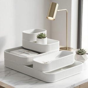 Op de afbeelding: Een set witte, geribbelde, rechthoekige opbergbakjes gestapeld in een getrapte opstelling. De bakjes bevatten een klein wit notitieboekje met een pen, een kleine vetplant in een potje en een paar metalen paperclips. Een gouden bureaulamp staat ernaast.