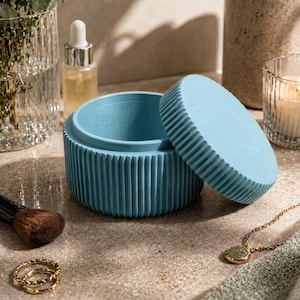 May include: A light blue cylindrical container with a ribbed exterior texture and a matching circular lid resting beside it. The container features a smooth interior basin and a matte finish, designed for holding small items on a flat surface.