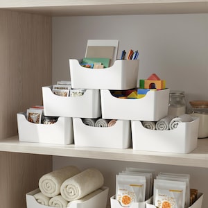 May include: A set of white plastic storage bins with open-front designs, stacked on shelves. The bins contain various items including notebooks, pens, colourful building blocks, rolled grey towels, and several small food pouches with printed labels.