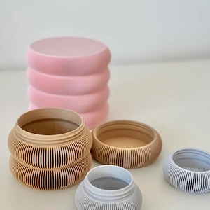 May include: A collection of cylindrical containers with ribbed, textured sides. One tall pink container with a lid, one medium tan container with a lid, one small tan base, one small grey container, and one small grey base are arranged on a flat surface.