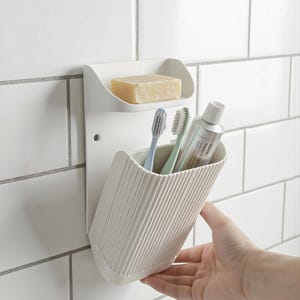 May include: A white plastic bathroom organiser fixed to white tiles. It features a top shelf with a bar of soap and a lower, ribbed section holding toothbrushes and toothpaste. Designed to store bathroom essentials, it's a practical storage solution.