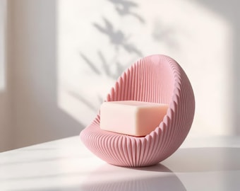 May include: A pale pink soap bar rests inside a rounded, shell-shaped soap dish. The dish features a textured, ribbed exterior with a curved, semi-circular design that cradles the rectangular soap bar securely within its centre.