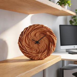 May include: A circular wall clock featuring a textured, terracotta-coloured face with a swirling, fabric-like pattern. The clock has two thin, black hands positioned at the centre, contrasting against the matte, rippled surface of the round dial.