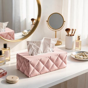 May include: A rectangular tissue box cover featuring a soft, light pink quilted exterior with a diamond-stitched pattern. The cover is designed to hold a standard stack of white facial tissues, which are visible protruding from the top opening.