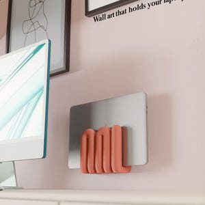 Op de afbeelding: Een koraalkleurige, golvende wandhouder die een zilveren laptop vasthoudt. De houder heeft een reeks afgeronde, verticale lussen die het apparaat tegen de muur bevestigen. De tekst "Wall art that holds your laptop" is zichtbaar boven de houder.