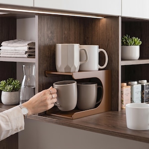 Può includere: Un organizer per scaffali in metallo marrone a forma di C che contiene due tazze sul ripiano inferiore e due tazze su quello superiore. L'organizer è posizionato su uno scaffale in legno scuro con altri oggetti da cucina e piccole piante grasse.