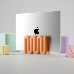 May include: A silver laptop with a black Apple logo is held upright by a matte peach-coloured, wavy, serpentine-shaped stand. The stand features a continuous, repeating S-curve design that provides a stable base for the electronic device.
