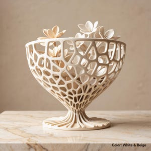 May include: A white and beige bowl with a complex, open-work lattice structure resembling a honeycomb or organic web. The bowl features a wide, rounded top that tapers down to a narrow stem and a circular, ribbed base, filled with several light-coloured flowers.