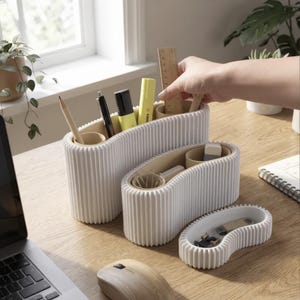Puede incluir: Un juego de tres organizadores de escritorio blancos, acanalados y con forma de riñón con interiores color canela. El recipiente más grande contiene un lápiz, un bolígrafo, un resaltador y una regla de madera. El mediano contiene objetos y el pequeño clips.