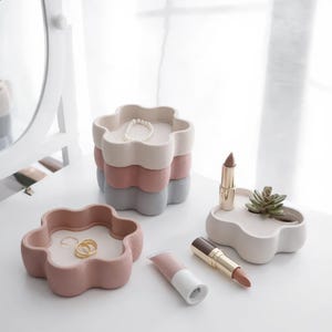Könnte beinhalten: Ein Stapel aus drei blumenförmigen Schalen in Creme, Altrosa und Grau, daneben eine einzelne altrosa Schale mit goldenen Ringen, eine cremefarbene Schale mit einer kleinen Sukkulente und zwei Lippenstifte. Die Schalen haben eine matte Oberfläche.