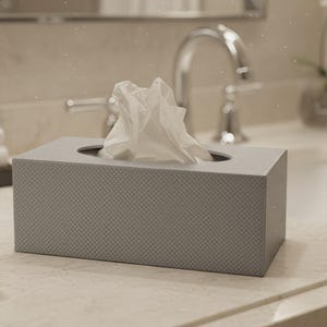 Puede incluir: Una caja rectangular para pañuelos con un acabado texturizado en gris claro. La caja presenta una abertura ovalada central en la parte superior, a través de la cual se extrae un pañuelo de papel blanco que sobresale ligeramente de la superficie.