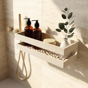 May include: A rectangular beige shower caddy with a textured, slatted front and a decorative cutout base. It holds two amber-coloured pump bottles, a round beige scrub brush, and a small white vase containing a green eucalyptus branch.