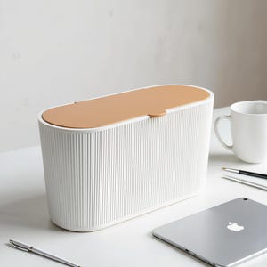 Puede incluir: Un contenedor de almacenamiento de forma ovalada con un exterior blanco acanalado y una tapa de color tostado sólido. El contenedor presenta un diseño de tapa abatible con una pequeña pestaña para abrirlo, ofreciendo una apariencia estructurada.