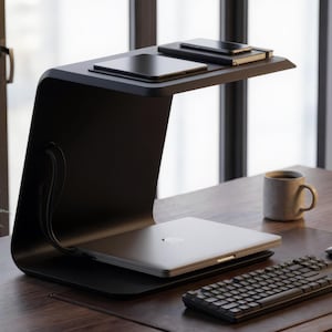 Pode incluir: Um organizador de secretária preto mate em forma de C com uma prateleira inferior que suporta um portátil prateado fechado e uma prateleira superior com um tablet preto e um caderno preto com um smartphone em cima, com cabos pretos laterais.
