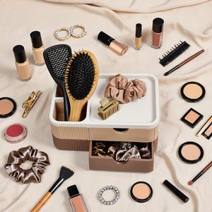 May include: A multi-tiered plastic desk organiser in shades of cream and brown, featuring a drawer and open compartments. It is surrounded by various beauty items including hairbrushes, scrunchies, makeup brushes, compacts, and bottles.
