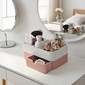 Può includere: Un organizer da scrivania a due livelli con una sezione superiore bianca a coste con scomparti multipli e un cassetto inferiore rosa a coste. L'organizer contiene vari articoli cosmetici, tra cui flaconi, pennelli e un elastico in raso elastico per capelli.