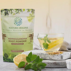 Op de afbeelding: Een groene en witte zak "Nature Origins Ashwagandha Root Powder" staat naast een glazen theekopje met citroen en munt. Een halve citroen en groene bladeren staan op de voorgrond. De zak vermeldt "Food Supplement" en "Premium Quality".