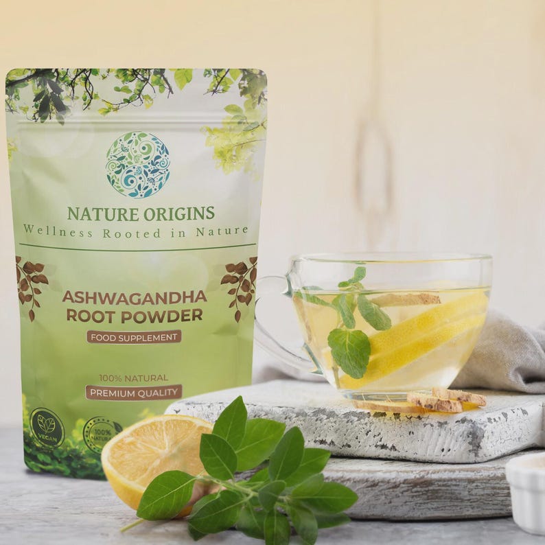 Op de afbeelding: Een groene en witte zak "Nature Origins Ashwagandha Root Powder" staat naast een glazen theekopje met citroen en munt. Een halve citroen en groene bladeren staan op de voorgrond. De zak vermeldt "Food Supplement" en "Premium Quality".