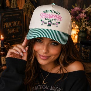 May include: A cream-colored baseball cap with a green and white striped brim. The front features embroidered text that reads Midnight Margarita Society Est. 1998, accompanied by a green blender icon and two pink margarita glass illustrations.
