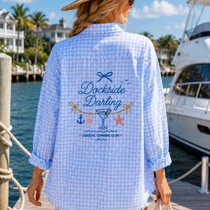 May include: A light blue and white gingham button-down shirt featuring a back graphic with the text "Dockside Darling" and "COASTAL COWGIRL CLUB," alongside illustrations of a bow, a martini glass, an anchor, a starfish, and a diamond shape.