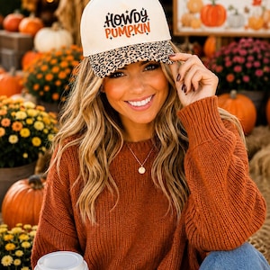 May include: A cream-colored baseball cap featuring the text "Howdy PUMPKIN" in brown and orange lettering with small star accents. The brim of the cap is decorated with a brown and black leopard print pattern, and the cap has a button on top.