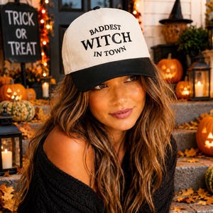 May include: A baseball cap with a cream-colored crown and a black brim. The front of the cap features the black embroidered text "BADDEST WITCH IN TOWN" in a mix of serif and sans-serif fonts. The cap has a small black button on the top center.