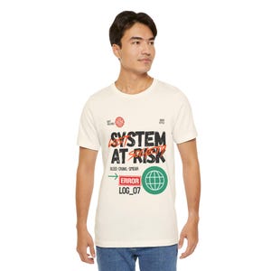 May include: A cream-colored short-sleeve t-shirt featuring a graphic print with the text SYSTEM AT RISK, LOST SOCIETY, BLEED-CRAWL-SMEAR, ERROR, and LOG_07. The design includes a green globe icon, a red rectangular border, and a green arrow.