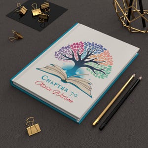 May include: A white hardback notebook with a teal spine featuring a graphic of an open book with a colourful tree growing from its pages. The text reads "CHAPTER 70" in teal and "Olivia Wilson" in red script. Two pencils lie beside the notebook.