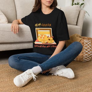 Könnte beinhalten: Schwarzes T-Shirt mit einer Grafik einer Katze, die auf einem Laptop-Bildschirm schläft. Der Text lautet "404 ERROR" und "MOTIVATION NOT FOUND". Die Katze ist gelb und orange. Die Person trägt blaue Jeans und weiße Turnschuhe.