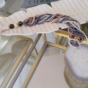 May include: A beaded necklace with a large agate pendant, displayed on a white, leaf-shaped dish. The necklace has a tassel and is made of beads in shades of grey, pink, and white. The agate pendant is approximately 10 cm in diameter.