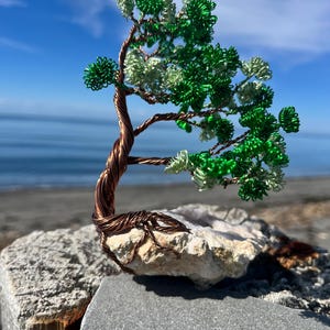Peut inclure: Une sculpture en fil de fer représentant un arbre avec un tronc et des branches torsadés de couleur cuivre. Le feuillage est composé de fils enroulés verts et vert clair. L'arbre est fixé sur une base en pierre brute et claire.