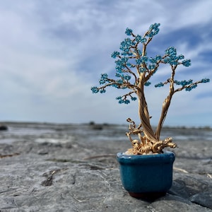 May include: A miniature bonsai tree sculpture with a gold-colored wire trunk and branches. The tree's leaves are made of small, light blue beads. The bonsai is planted in a small, round, blue ceramic pot. The background features a cloudy sky and a rocky surface.